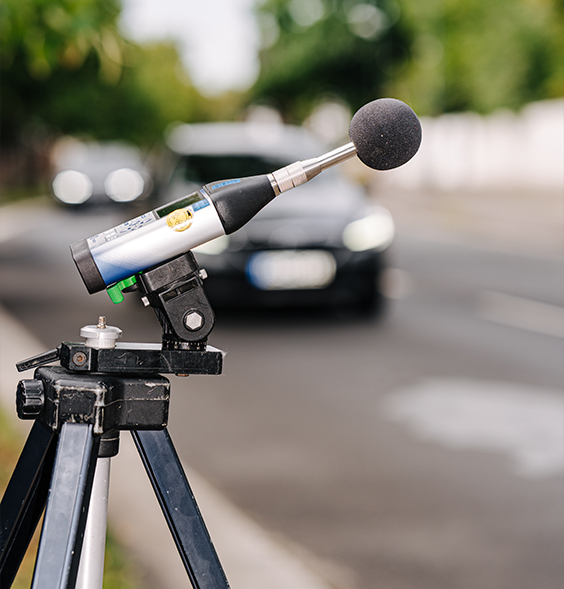 Ein Schallpegelmesser auf einem Stativ misst den Verkehrslärm an einer Straße.