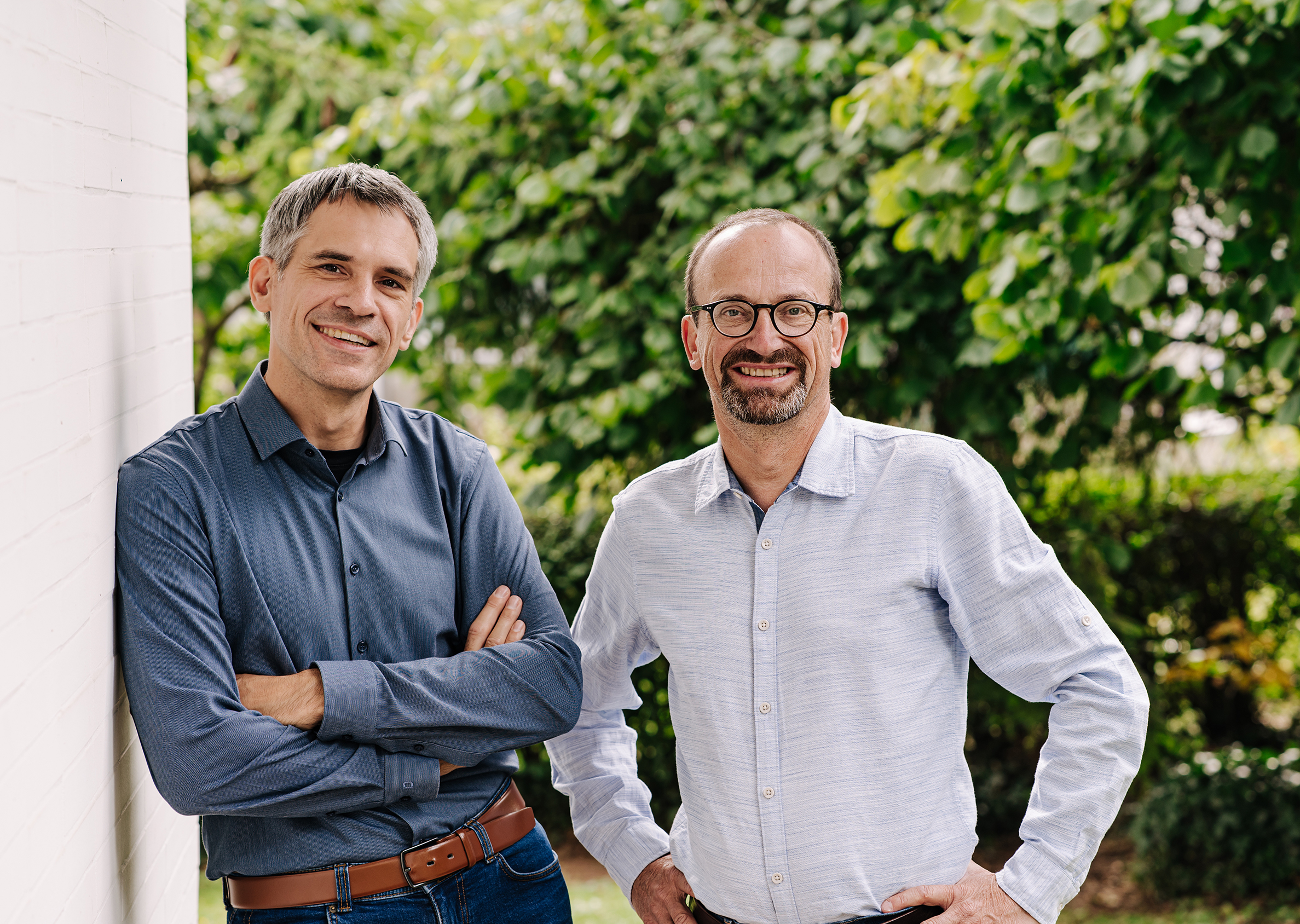Zwei Mitarbeiter stehen entspannt im Grünen und lächeln in die Kamera.