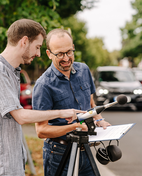 Zwei Mitarbeiter führen eine Schallmessung im Außenbereich durch und prüfen gemeinsam die Werte eines Schallpegelmessers.